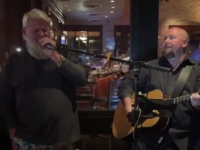 John Daly is singing on the mic as the other guy is playing the guitar.